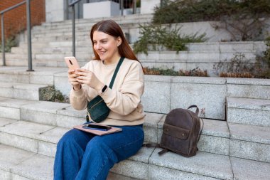 Açık havada akıllı telefon kullanan güzel, gülümseyen kız öğrenci. Şehir caddesinde merdivenlerde oturan neşeli genç bir kadın. Şehir yaşam tarzı konsepti. Gezgin, Çevrimiçi Eğitim veya Alışveriş