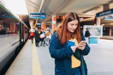Genç kızıl saçlı kadın arka plandaki elektrikli trende akıllı telefon kullanarak istasyon platformunda bekliyor. Demiryolu taşımacılığı konsepti, Gezgin