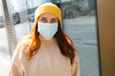 Yüzünde koruyucu maske olan sarı şapkalı neşeli genç bir kadın. Virüs yayılmış grip önleme karantinası.