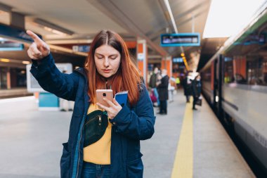 Sırt çantasıyla istasyonda bekleyen genç kızıl saçlı kadın akıllı telefon kullanarak arka plandaki treni işaret ediyor. Demiryolu taşımacılığı konsepti, Gezgin