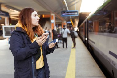 Genç kızıl saçlı kadın arka plandaki elektrikli trende akıllı telefon kullanarak istasyon platformunda bekliyor. Demiryolu taşımacılığı konsepti, Gezgin