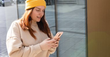 Şehir caddesinde yürüyen neşeli genç kadın akıllı telefonunu kontrol ediyor. Açık havada akıllı telefon kullanan sarı şapkalı güzel bir kadının portresi. Şehir yaşam tarzı konsepti. Gezgin