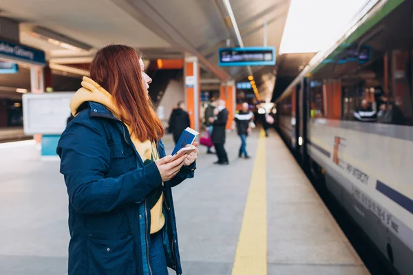 Mujer pelirroja joven esperando en la plataforma de la estación con mochila en el tren eléctrico de fondo utilizando el teléfono inteligente. Concepto de transporte ferroviario, Viajero — Foto de Stock