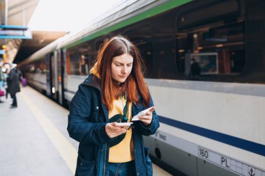 Genç kızıl saçlı kadın arka plandaki elektrikli trende akıllı telefon kullanarak istasyon platformunda bekliyor. Demiryolu taşımacılığı konsepti, Gezgin