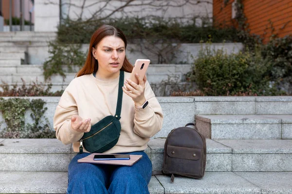 Mujer joven molesta usando el teléfono y leyendo malas noticias, chica pensativa triste sentado en las escaleras en la calle de la ciudad. Gente negativa emoción — Foto de Stock