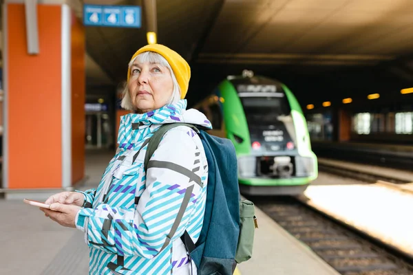 Vieja mujer feliz con mochila en el tren de espera sombrero amarillo en la plataforma de la estación y el uso de teléfono inteligente en el fondo urbano. Concepto de transporte ferroviario — Foto de Stock