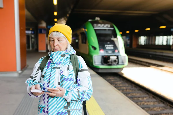 Vieja mujer feliz con mochila en sombrero amarillo comprobando su billete y utilizando el teléfono inteligente en el tren de fondo. Concepto de transporte ferroviario. Concepto de transporte ferroviario — Foto de Stock