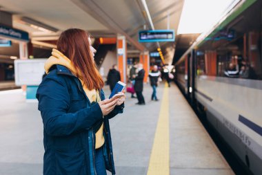 Genç kızıl saçlı kadın arka plandaki elektrikli trende akıllı telefon kullanarak istasyon platformunda bekliyor. Demiryolu taşımacılığı konsepti, Gezgin. Ukrayna 'da İstila.