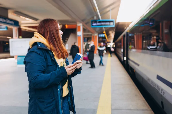 Mujer pelirroja joven esperando en la plataforma de la estación con mochila en el tren eléctrico de fondo utilizando el teléfono inteligente. Concepto de transporte ferroviario, viajero. Invasión en Ucrania. — Foto de Stock