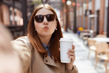 Moda ofis kıyafetleri içinde neşeli, genç, kızıl saçlı bir kadın ve güneş gözlüklü bir fincan çay eşliğinde dışarıda poz veriyor. Mockup, bir kadın selfie çekiyor ve elinde biyo-kahve fincanı tutuyor. Kentsel yaşam tarzı konsepti, model