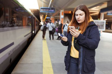 Genç kızıl saçlı kadın arka plandaki elektrikli trende akıllı telefon kullanarak istasyon platformunda bekliyor. Demiryolu taşımacılığı konsepti, Gezgin