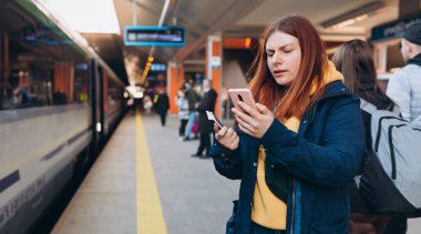 Genç kızıl saçlı kadın arka plandaki elektrikli trende akıllı telefon kullanarak istasyon platformunda bekliyor. Demiryolu taşımacılığı konsepti, Gezgin. Ukrayna 'da İstila.