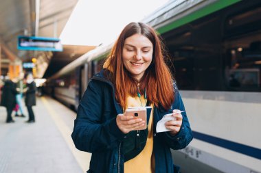 Genç kızıl saçlı kadın arka plandaki elektrikli trende akıllı telefon kullanarak istasyon platformunda bekliyor. Demiryolu taşımacılığı konsepti, Gezgin