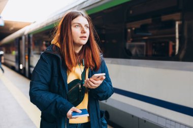 Arka plandaki tren istasyonunda bekleyen genç mülteci kızıl saçlı kadın kaybetmiş ve akıllı telefon kullanmış. Demiryolu taşımacılığı konsepti, Gezgin. Ukrayna 'da istila. Rus saldırısı