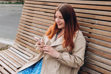 Mutlu, genç bir kadın bankta oturuyor ve şehir caddesinde akıllı telefon kullanıyor. Şehir yaşam tarzı konsepti. Gezgin, Online alışveriş, uygulama kullanarak