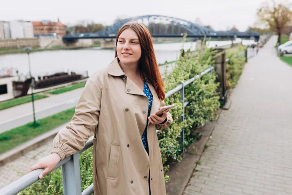 Güzel mutlu kızıl saçlı kadın akıllı bir telefon kullanıyor ve dışarıda gülümsüyor. Şehir yaşam tarzı konsepti. Şehir caddesinde yürüyen kız akıllı telefonunu kontrol ediyor. Bahar geldi.