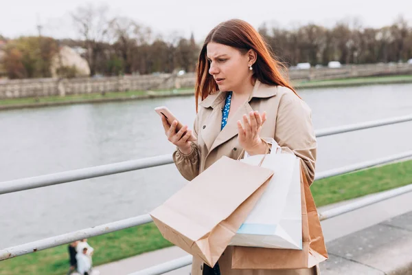 Upset young woman using phone and reading bad news, sad pensive girl walking on city street. Negative people emotion. Redhead girl is holding paper shopping bags, no special offer, the end of sales — Stock Photo, Image