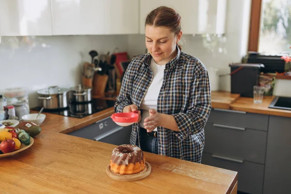 Happy Woman making homemade cake with easy recipe, sprinkling powdered sugar on top. Young redhead girl pastry chef making chocolate cake in the kitchen. Baking Concept — стоковое фото