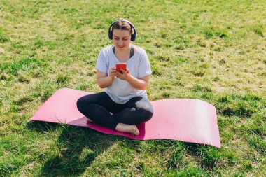 Güzel, genç, kızıl saçlı bir kadın antrenmandan sonra Yoga Mat 'te Smartphone ile dinleniyor. Kulaklıklı, müzik dinleyen ya da internete göz atan mutlu bir kız. Spor ya da yoga konsepti