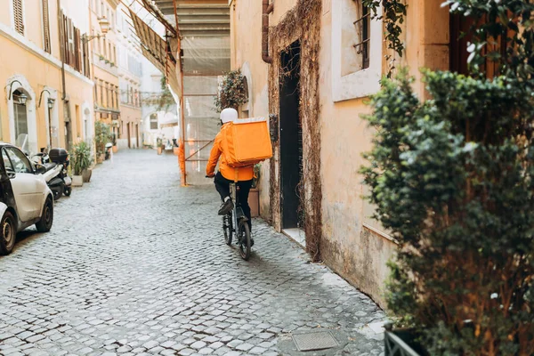 Young man delivering goods with a orange thermal backpack, riding a bicycle in the city. Courier on a bicycle delivering food. Quick shipping to customers — Fotografia de Stock