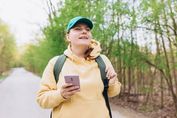 Portrét usměvavé krásné ženy v zelené čepici sms s telefonem na přírodním pozadí. Šťastná žena s batohem používá smartphone v parku venku, jaro. Cestovatel — Stock fotografie