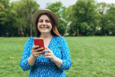 Şapkalı gülümseyen güzel bir insanın portresi. Doğa arka planında telefonuyla mesajlaşıyor. Sırt çantalı mutlu kadın dışarıda parkta akıllı telefon kullanıyor.