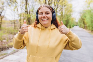 Kablosuz kulaklıkla müzik dinleyen ve doğa arka planında baş parmak gösteren mutlu bir genç kızın portresini yakından çek. Müzik aşığı müziğin tadını çıkarıyor. Yaşam tarzı kavramı.