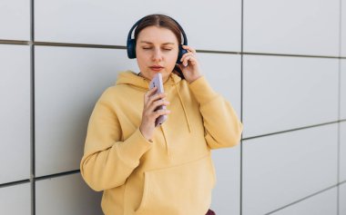 Kablosuz kulaklıkla müzik dinleyen ve gri arka planda şarkı söyleyen sevimli genç bir kızın yakın plan portresi. Müzik aşığı müziğin tadını çıkarıyor. Mobie cep telefonu diktafonundan kadın kayıt sesi