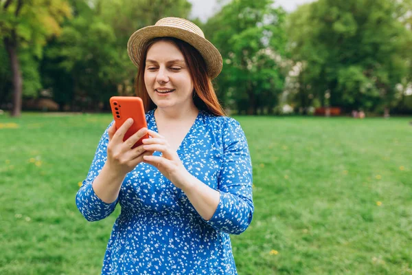 Şapkalı gülümseyen güzel bir insanın portresi. Doğa arka planında telefonuyla mesajlaşıyor. Mutlu kadın dışarıda parkta akıllı telefon kullanıyor.