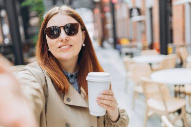 Moda ofis kıyafetleri içinde neşeli, genç, kızıl saçlı bir kadın ve güneş gözlüklü bir fincan çay eşliğinde dışarıda poz veriyor. Mockup, bir kadın selfie çekiyor ve elinde biyo-kahve fincanı tutuyor. Kentsel yaşam tarzı konsepti