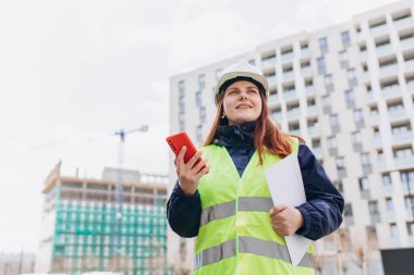 İnşaat alanındaki telefon ekranına bakan planlı bir mimar. Beyaz miğfer ve güvenlik yeleği giyen kadın müteahhit portresi.