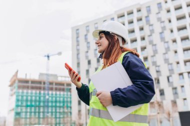 İnşaat alanında akıllı telefon kullanan, planlı bir mimar. Beyaz kask ve güvenlik yeleği giyen kadın müteahhit portresi, teknoloji konsepti.
