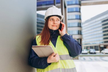 İnşaat alanında akıllı telefon kullanan, planlı bir mimar. Beyaz kask ve güvenlik yeleği giyen kadın müteahhit portresi, teknoloji konsepti.