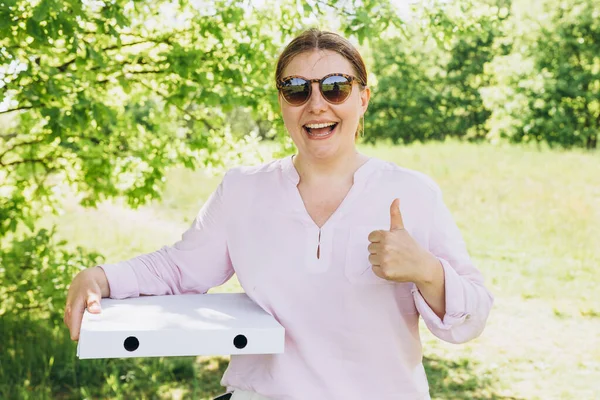 Happy Woman in sunglasses holding a box with pizza and showing thumbs up on nature background. Fast pizza delivery. Mock up. Fast food banner