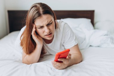 Üzgün genç bir kadın hasta ya da üzgün hissediyor modern cep telefonu cihazını kullanıyor, üzgün bir kadın yatak odasında dinleniyor akıllı telefonu tutuyor ve kötü haberleri okuyor