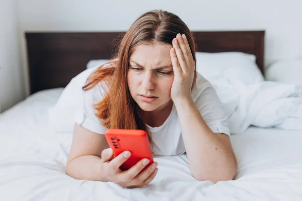 Upset young woman lying in bed feel sick or distressed using modern cellphone gadget, sad millennial female relax in bedroom hold smartphone read bad news