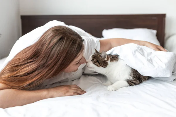 Happy young woman with cat in bed at home. In cold weather, the pet warms up under a blanket. Pet friendly and grooming concept. Stray kitten sleep on bed. Cozy home background