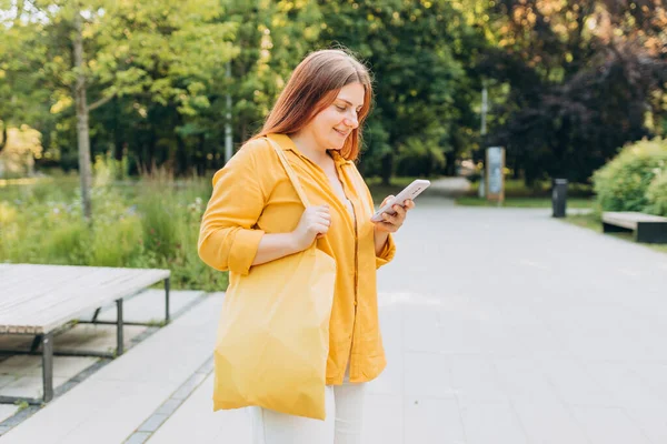 Stylish happy woman walking with yellow cotton bag in the city on summer day and holding smart phone. Urban lifestyle concept. Check social networks. Environmental Conservation Recycling mockup.