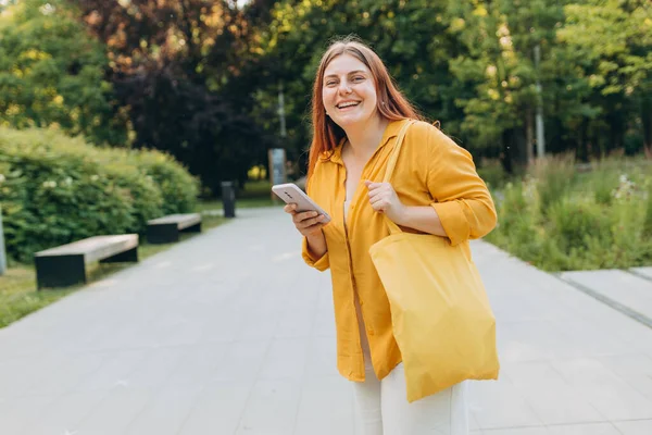 Beautiful woman walking with yellow cotton bag in the city on summer day and holding smart phone. Urban lifestyle concept. Check social networks. Environmental Conservation Recycling mockup.