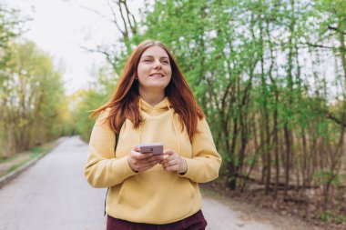 Gülümseyen kızıl saçlı güzel bir kadının portresi. Doğa arka planında telefonuyla SMS yolluyor. Mutlu kadın, bahar zamanı parkın dışında akıllı telefon kullanıyor. Gezgin