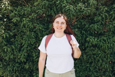 Sırt çantalı, beyaz tişörtlü ve yeşil doğa duvarına poz veren hippi kadın. Boş t-shirt, bitki arkaplanı, metin veya tasarım için boş yer.