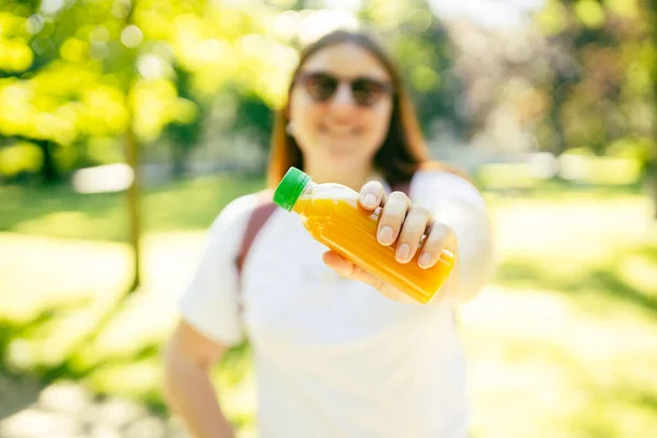 Young stylish woman with juice standing over nature background. Redhead prety girl walking in the park and holding a bottle of fresh cold smoothie in a warm sunny day.