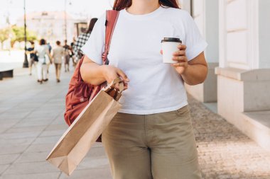 Beautiful fashionable young woman in sunglasses with shopping paper bag standing on city street, black friday. Urban lifestyle concept. Attractive female drinking a coffee in paper bio cup.