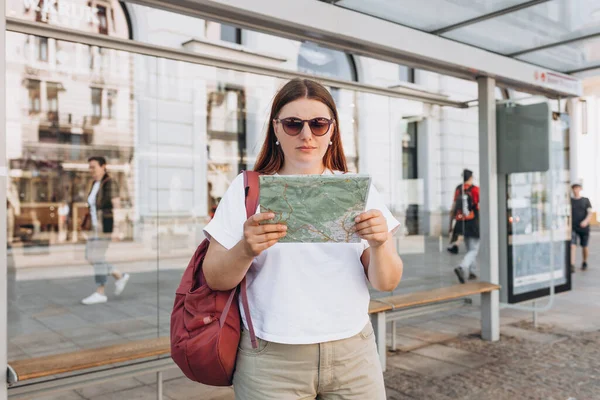 A cheerful caucasian redhead woman of an urban bus stop and using paper map. Urban travel and transportation concept. Girl search location