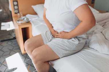 Young woman suffering from strong abdominal pain, casual style indoor shoot. Female sitting on bed in morning