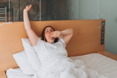 Beautiful redhead Woman waking up in the morning sitting in bed yawning. Good morning. Millennial Caucasian girl wake up in cozy white bed at home