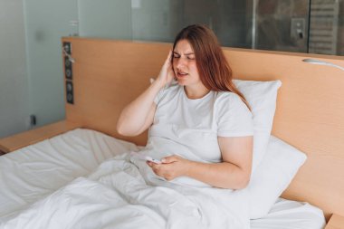 Upset young woman lying in bed feel sick or distressed using cellphone gadget. Sad millennial female relax in bedroom, hold smartphone and read bad news. Women having headache, hands on temples