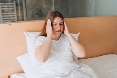 Young Woman with hard headache holding hands on head. Covid-19 new variant - Omicron. Pandemic infection. Upset woman lying in bed feel sick or distressed