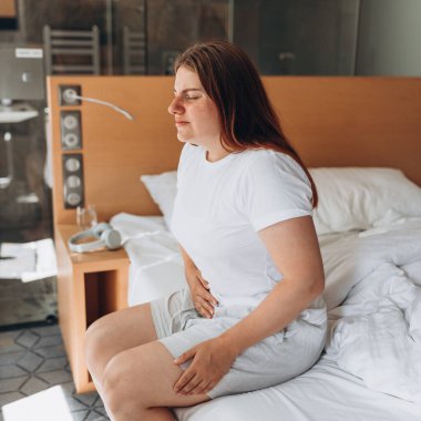 Young woman suffering from strong abdominal pain, casual style indoor shoot. Female sitting on bed in morning