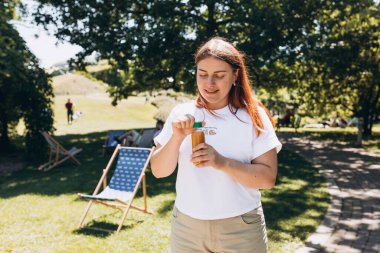 Güneş gözlüklü, dışarıda taze meyve suyu içen güzel bir kadın. Kızıl saçlı kız parkta yürüyor ve sıcak güneşli bir günde elinde koca bir şişe taze soğuk smoothie tutuyor..